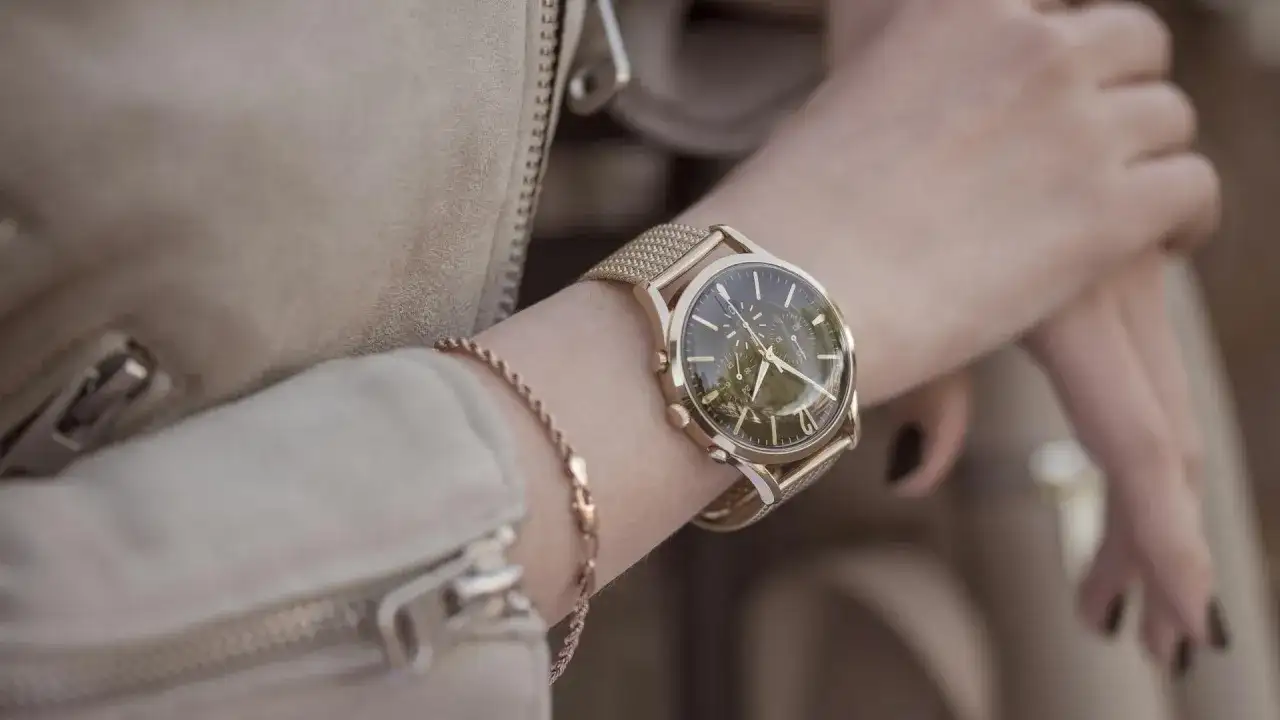 Damski zegarek na złotej bransolecie z ciemną tarczą, noszony na nadgarstku.