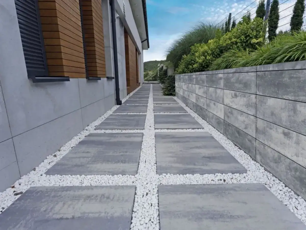 Nowoczesna ścieżka z prostokątnych płyt, tworząca opaskę z kostki brukowej, prowadzi wzdłuż domu i muru oporowego.