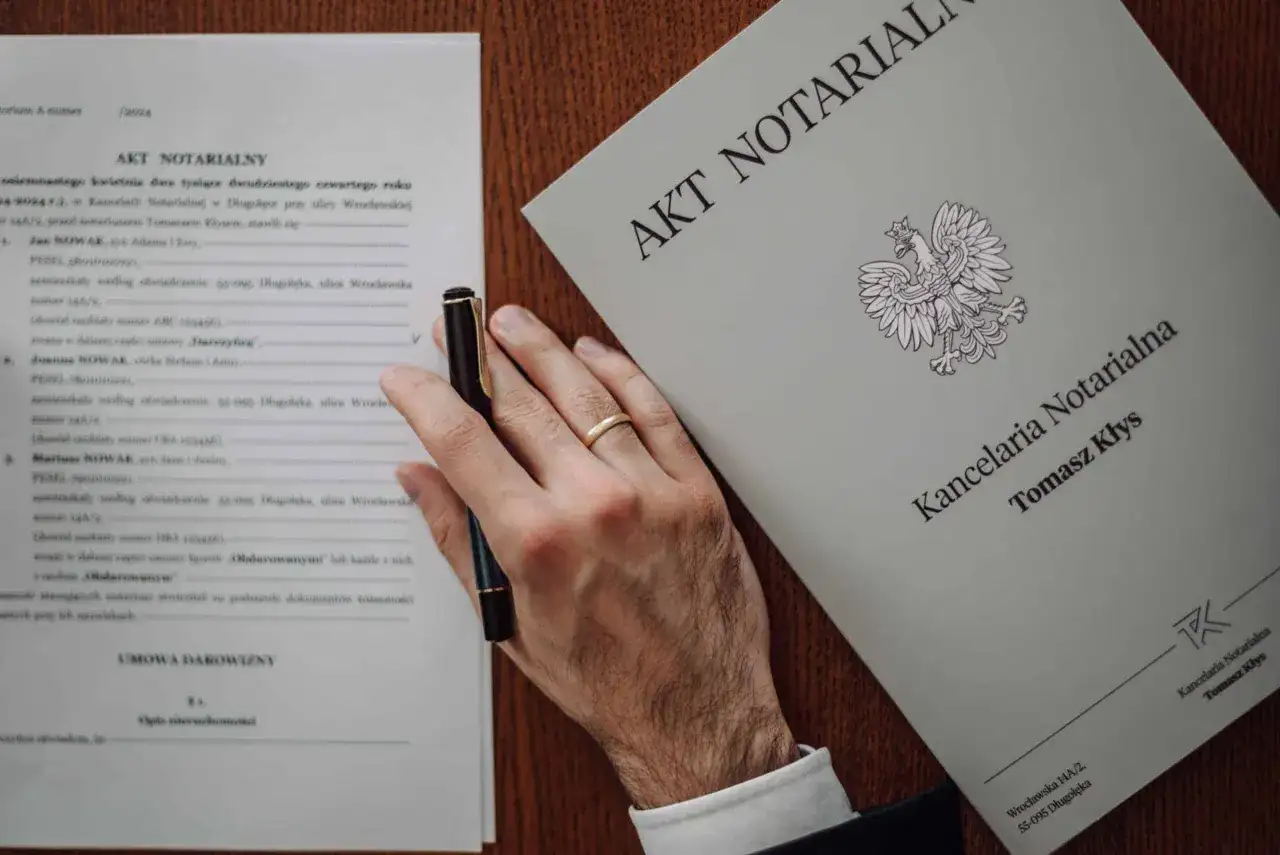 Dłoń z obrączką trzyma długopis nad dokumentem "Akt Notarialny". Zastanawiasz się, ile kosztuje poświadczenie podpisu u notariusza?