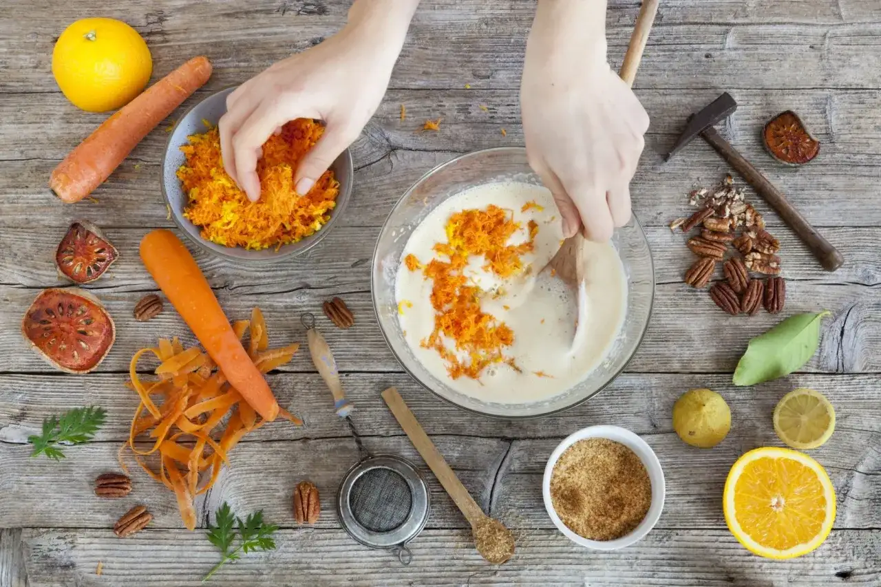 Ręce dodają startą marchewkę do ciasta. Czy gotowana marchewka jest zdrowa? Składniki na ciasto marchewkowe: marchew, pomarańcza, orzechy.