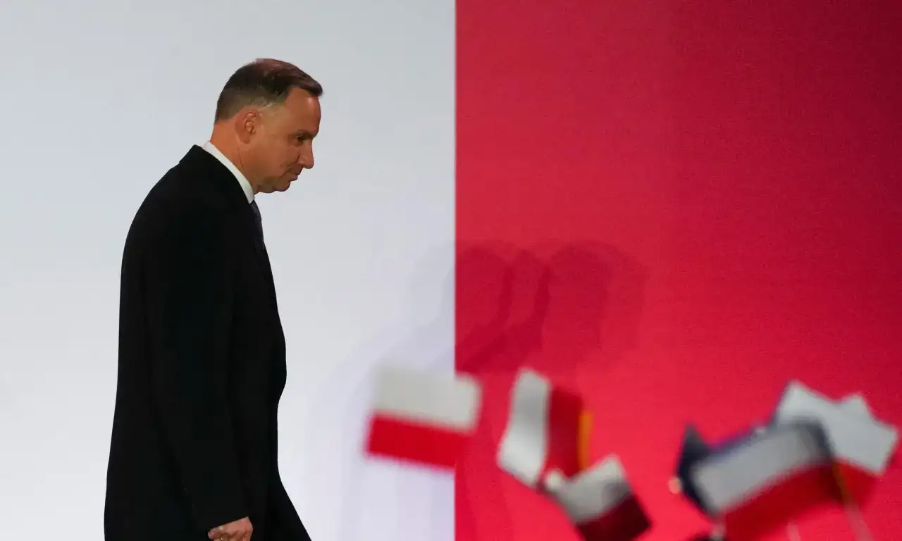 Andrzej Duda w czarnym garniturze, pochylony, idzie na tle czerwonej ściany z flagami. Czy to prawdziwe powody dla których Andrzej Duda zapowiedział serię wet do ustaw budżetowych?