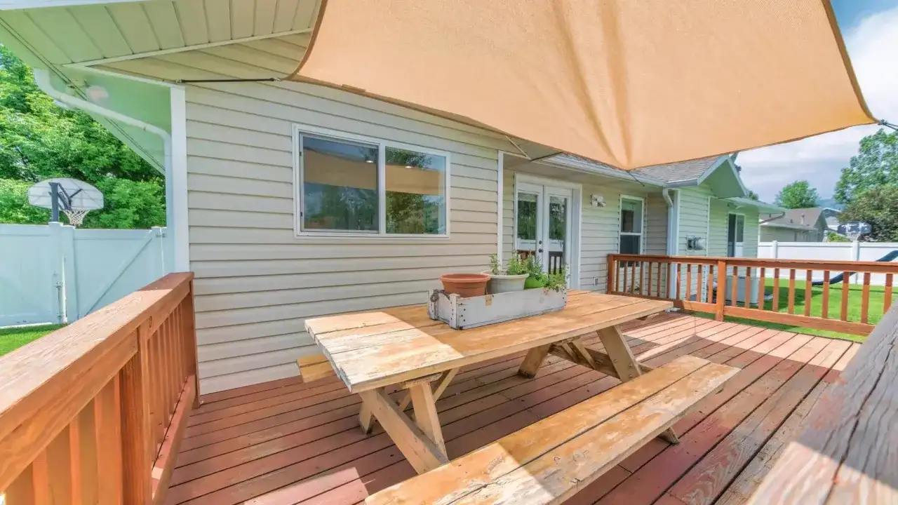 Drewniany stół piknikowy na tarasie pod beżowym żaglem cieniującym.