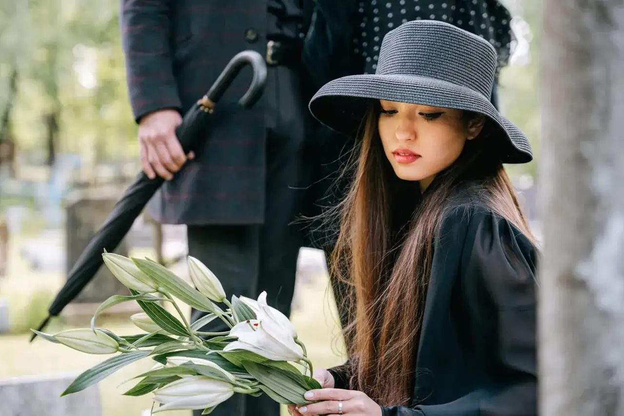 Smutna kobieta w kapeluszu i czarnej sukni trzyma białe lilie. Jej fryzura na pogrzeb jest schludna, a obok stoi mężczyzna z parasolem.