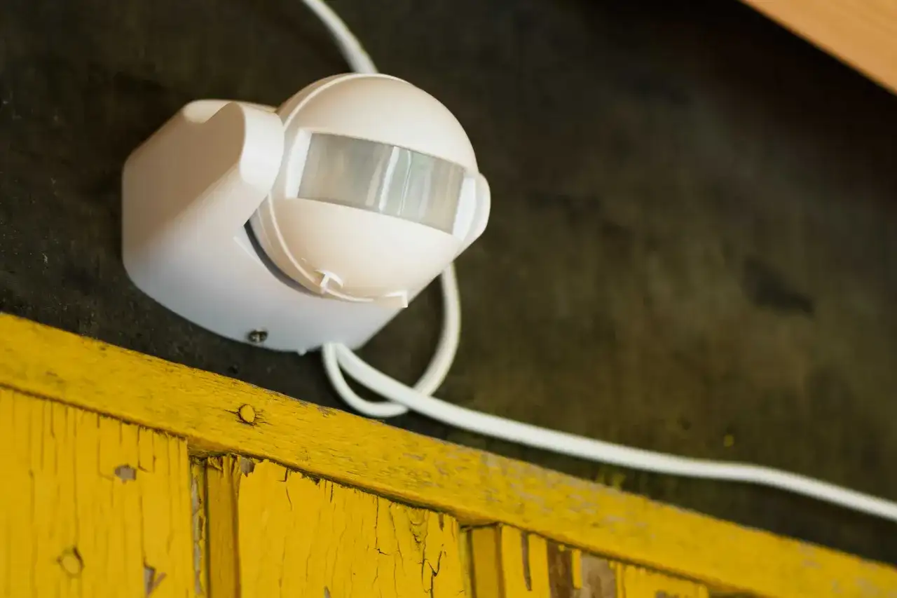 Biały czujnik ruchu na ciemnej ścianie, obok żółtej, pękającej deski. Dowiedz się, jak zmniejszyć czułość czujnika ruchu, by uniknąć fałszywych alarmów.