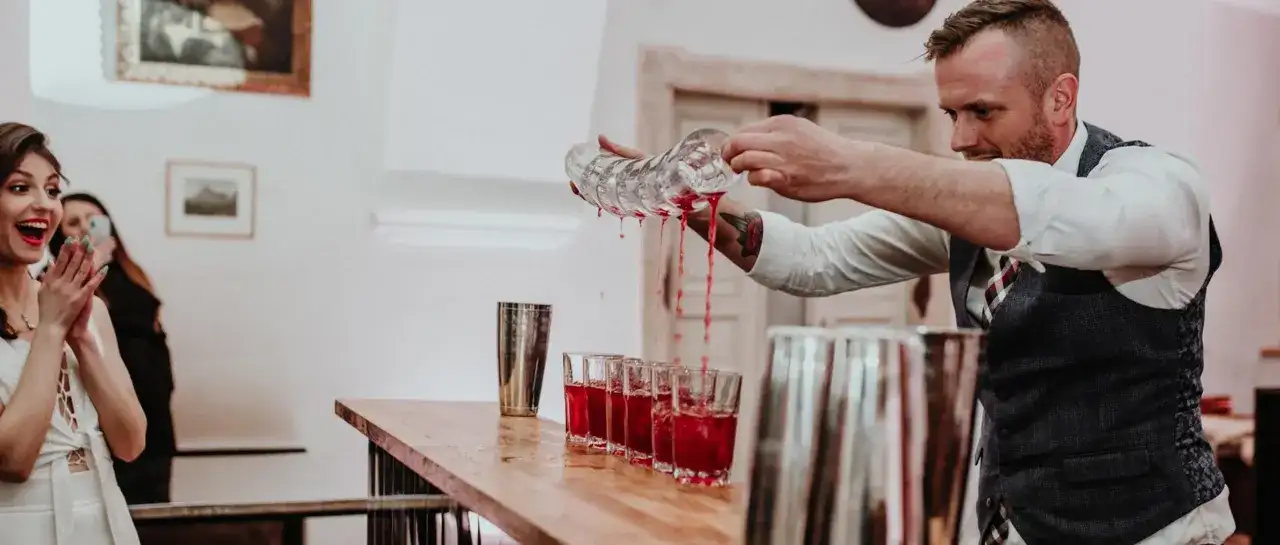 Barman nalewa czerwony napój do kieliszków na weselu, obok podekscytowana panna młoda.