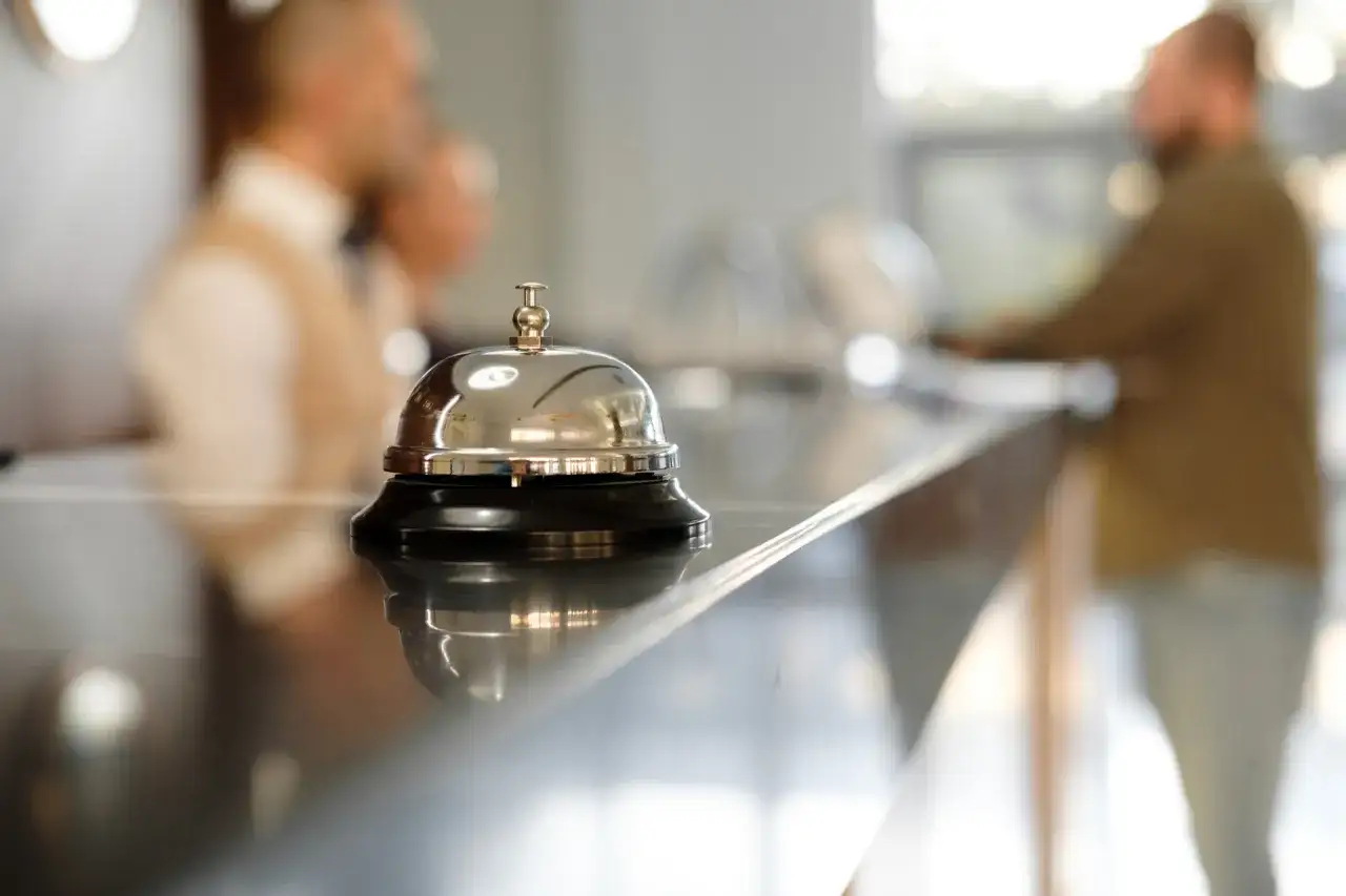 Dzwonek hotelowy na ladzie, symbol usług uzupełniających. W tle niewyraźne postacie personelu.