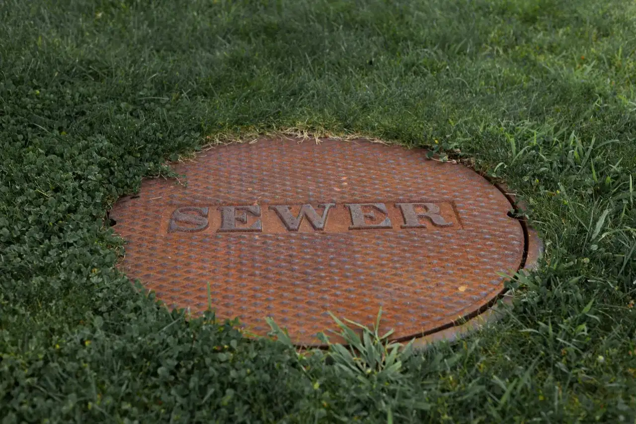 Zardzewiała pokrywa szamba z napisem "SEWER" w trawie. Czy na szambo trzeba mieć pozwolenie?