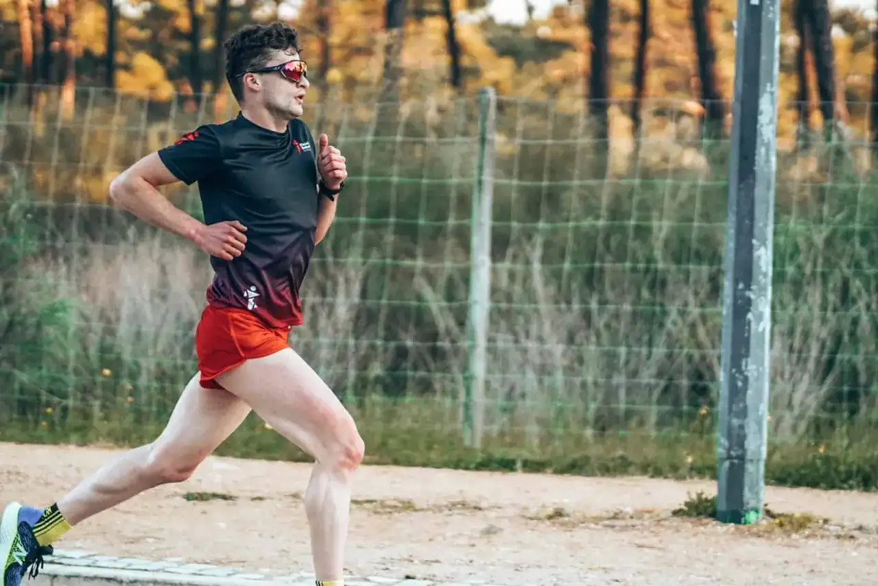 Mężczyzna w sportowym stroju biegnie, ćwicząc jak zwiekszyc kadencje w bieganiu. W tle siatka i drzewa.