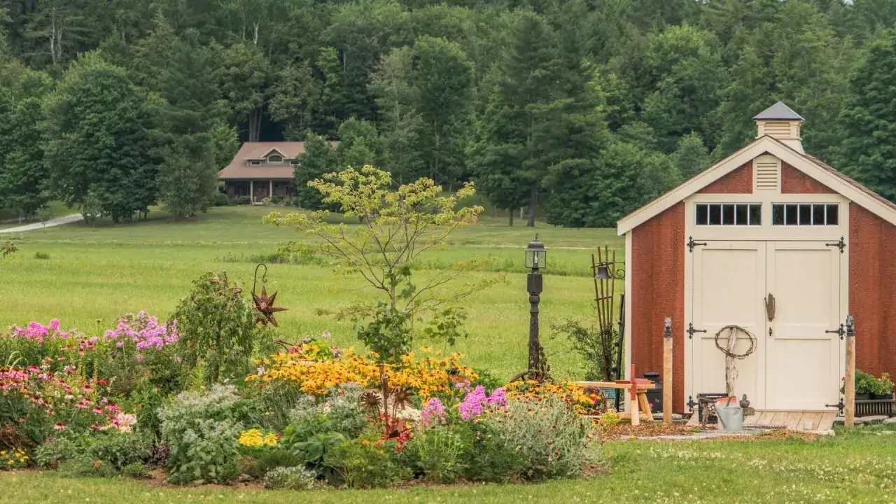Czerwony budynek gospodarczy definicja: schowek na narzędzia ogrodowe, otoczony kwitnącym ogrodem, z domem w tle.