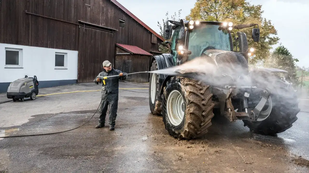 Mężczyzna myje traktor myjką gorącowodną przed stodołą.