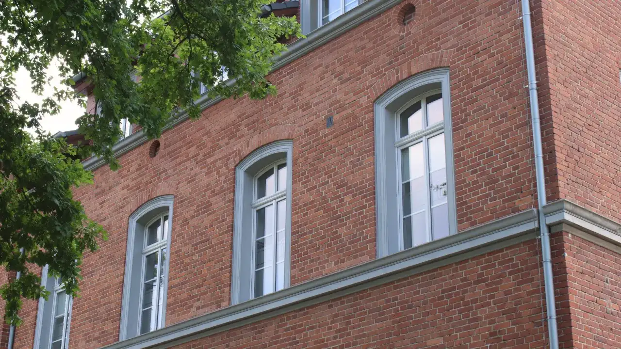 Eleganckie okna drewniane zabytkowe w ceglanej kamienicy, otoczone zielenią drzew.