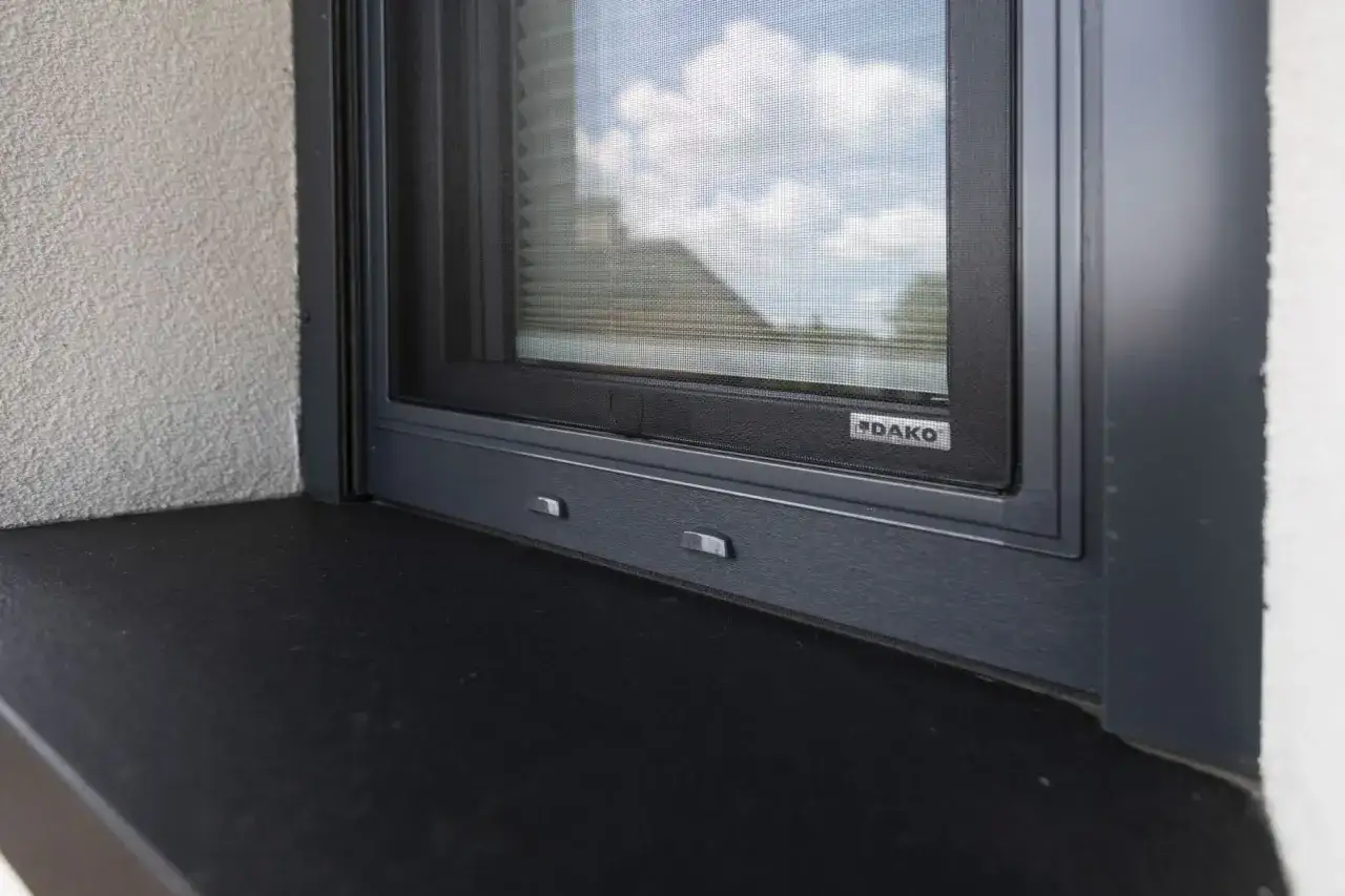 Montaż parapetów zewnętrznych do czoła okna, z widocznym logo DAKO. Ciemny parapet i szara rama okna.