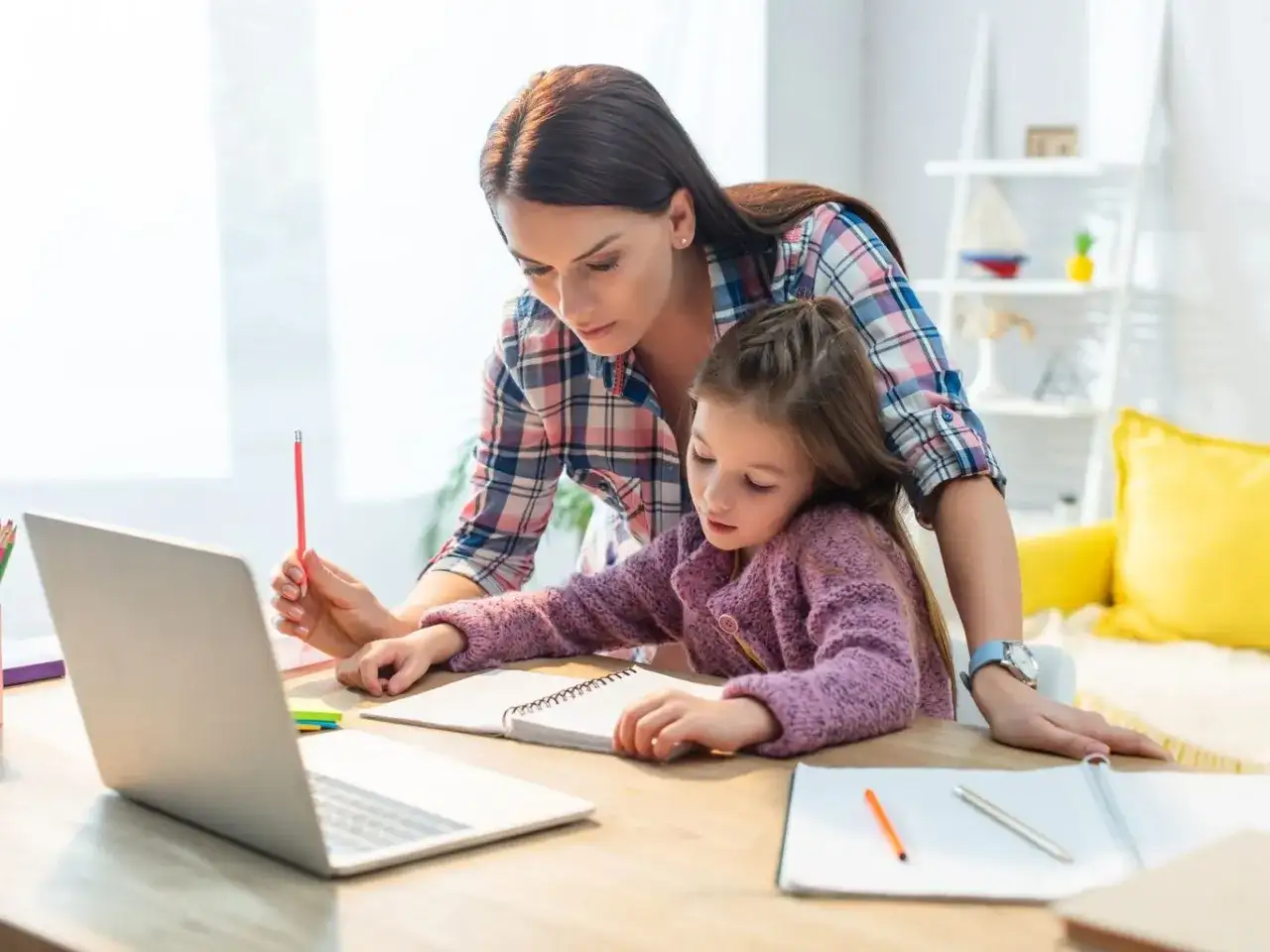 Kobieta pomaga dziecku w nauce matematyki przy biurku z laptopem i zeszytami.