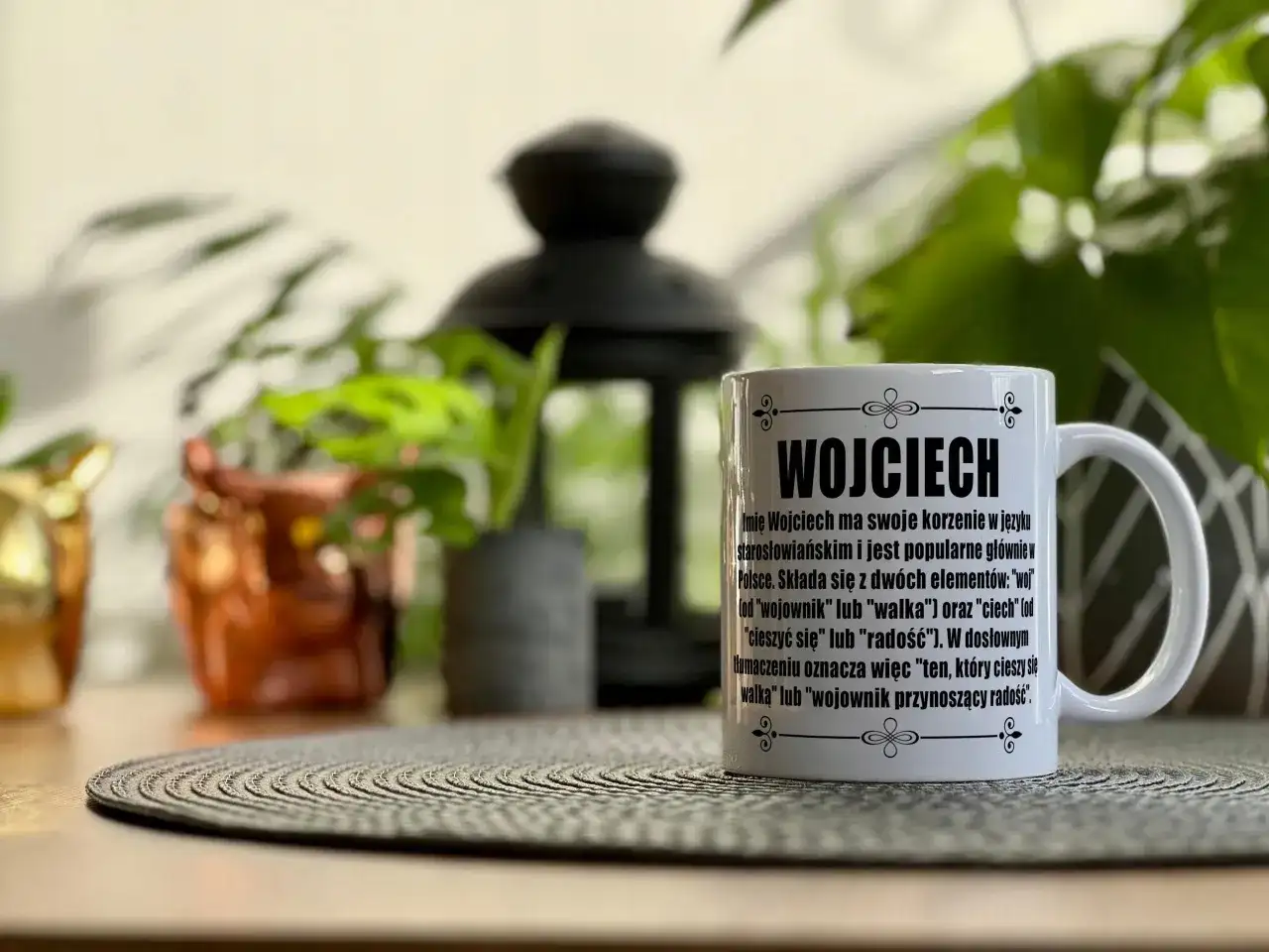 Biały kubek z napisem "Wojciech" i opisem znaczenia imienia, na tle roślin.