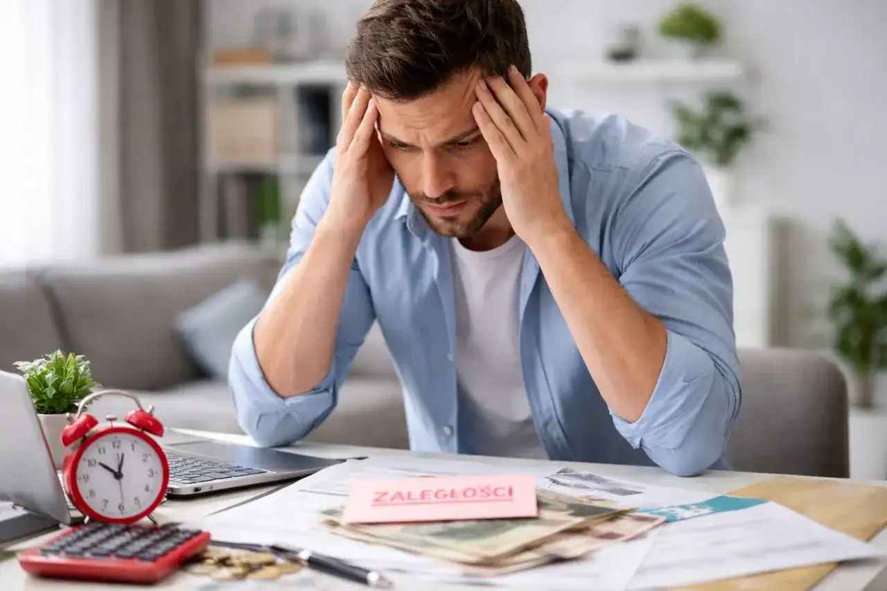 Zmartwiony mężczyzna z rękami na skroniach, przed nim dokumenty i karteczka "ZALEGŁOŚCI". Problem z niezapłaceniem składki ubezpieczeniowej w terminie.