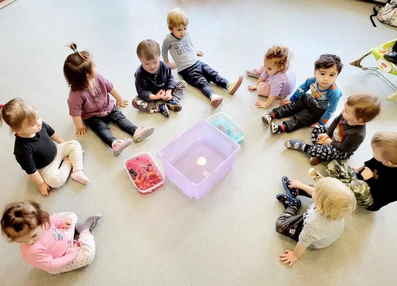 Dzieci siedzą w kółku na podłodze wokół pojemników z wodą i zabawkami.