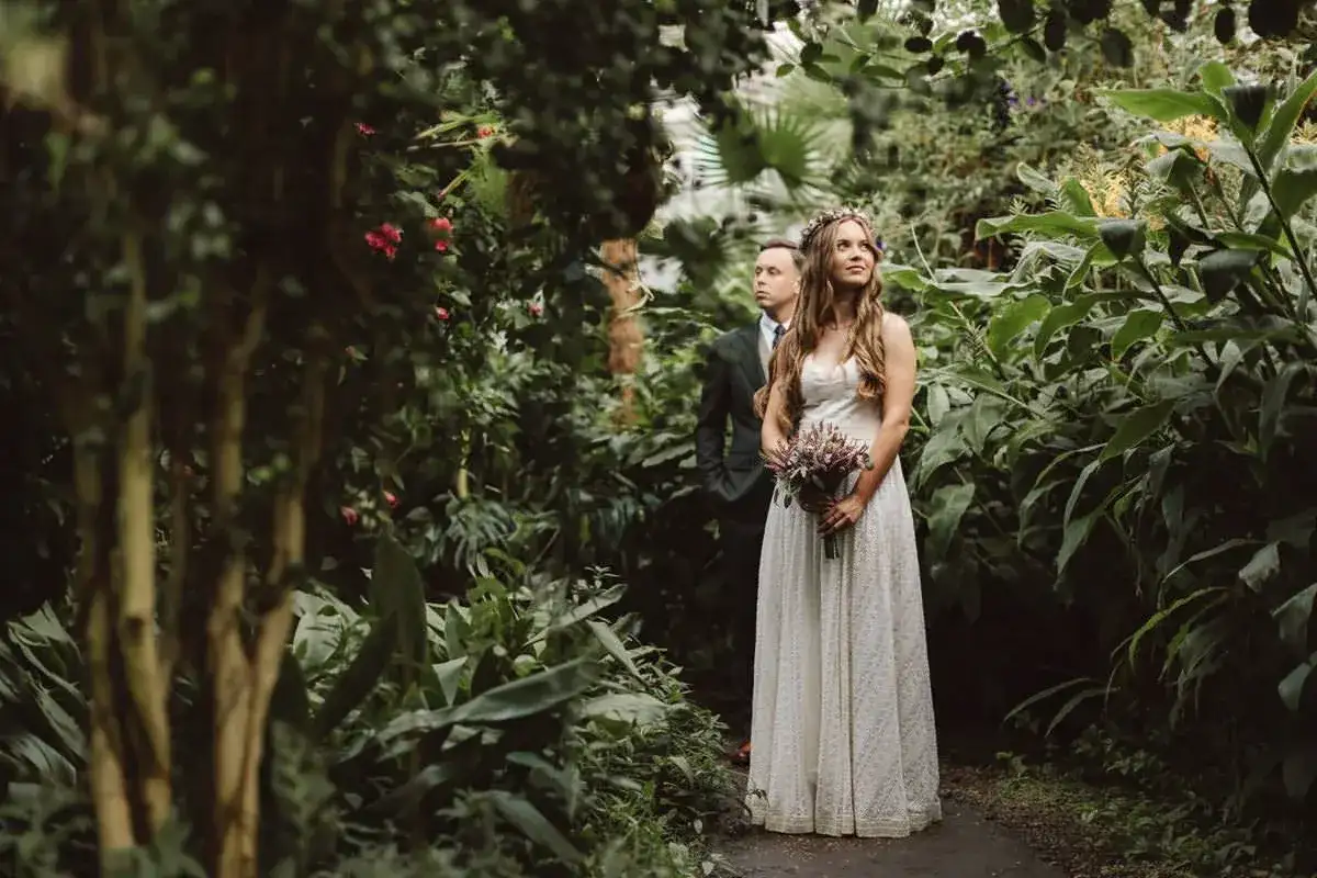Para dowodowa w otoczeniu bujnej zieleni. Panna młoda w białej sukni i wianku, pan młody w garniturze. Inspiracja, jak wybrać fotografa ślubnego.