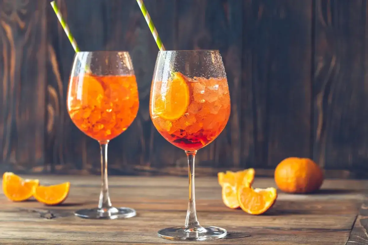 Dwa kieliszki Aperol Spritz z plasterkami pomarańczy i słomkami na drewnianym stole.