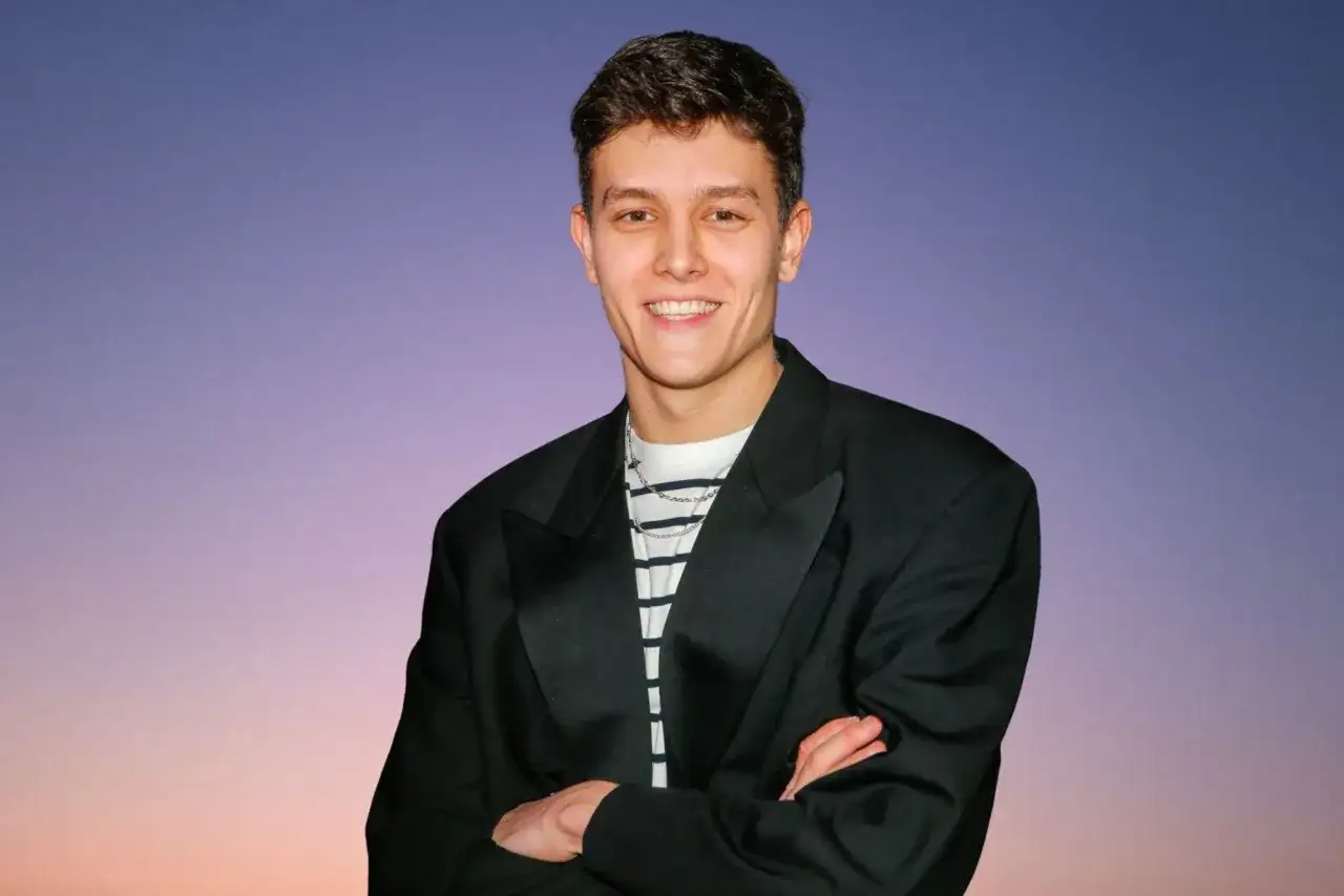 Młody mężczyzna w czarnej marynarce i pasiastym t-shircie z łańcuszkiem na szyi, na tle nieba.