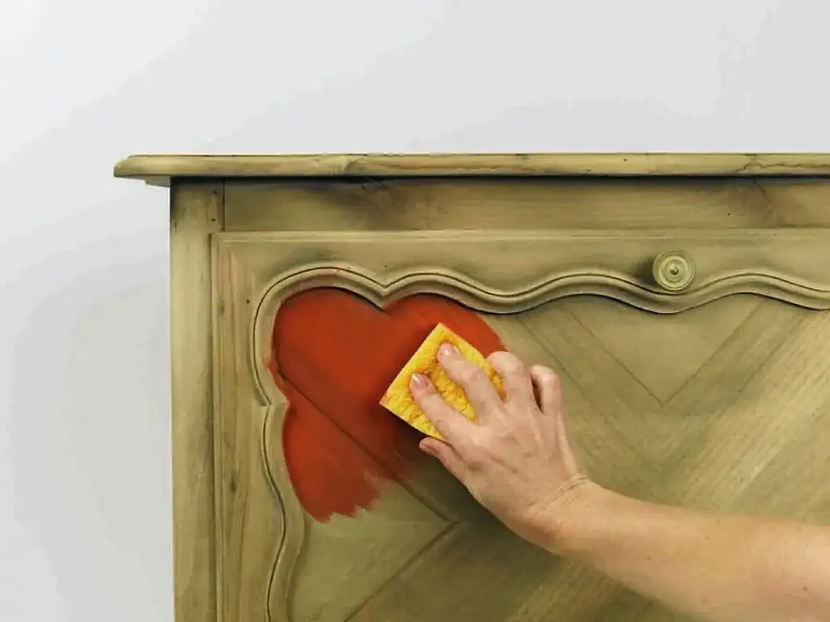 Dłoń z gąbką nakłada pomarańczową farbę na drewniany mebel.