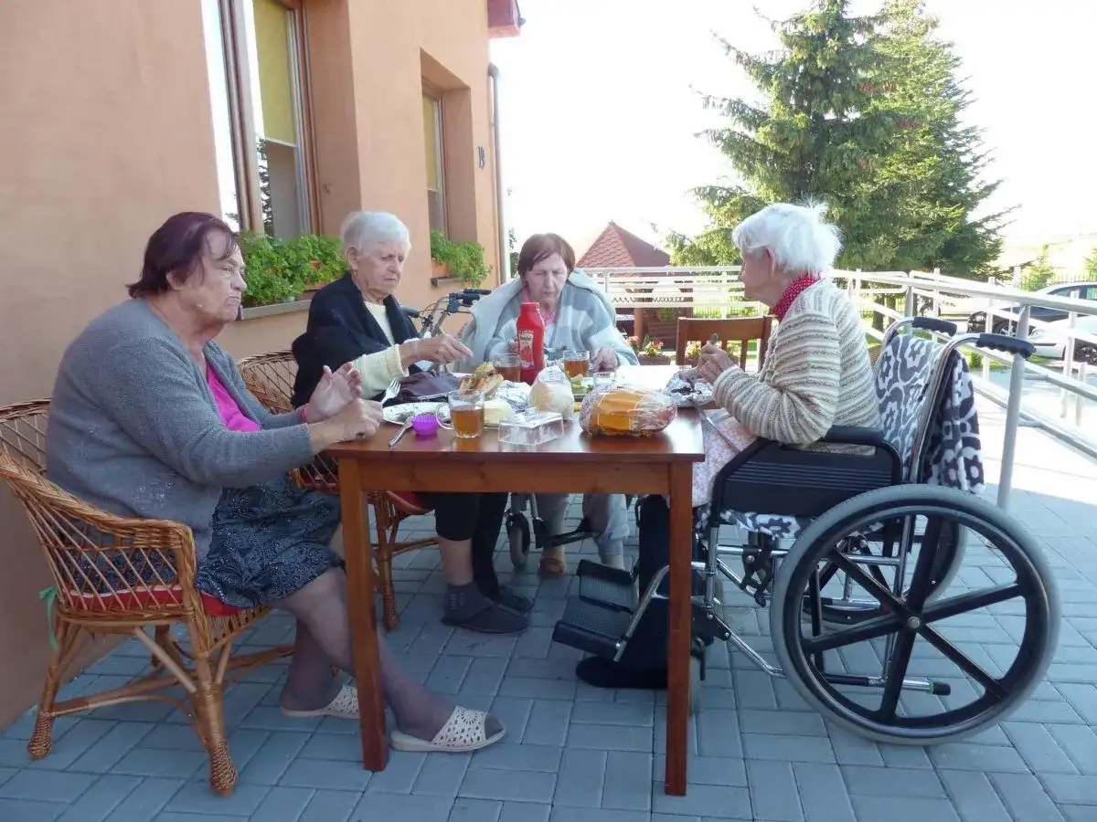 Cztery starsze osoby siedzą przy stole na zewnątrz, jedna na wózku inwalidzkim.