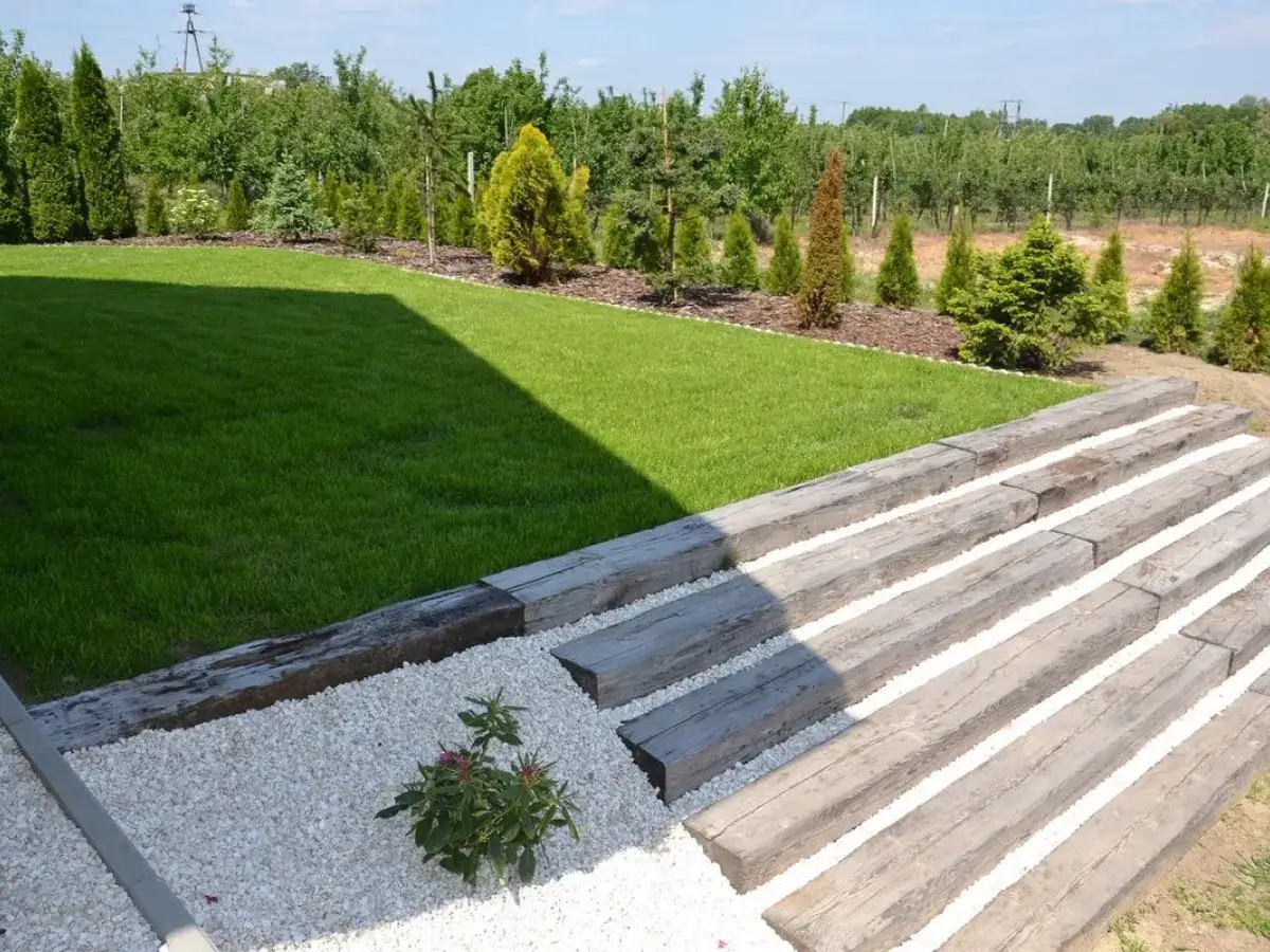 Schody na skarpie z podkładów kolejowych prowadzą do zadbanego ogrodu z trawnikiem i drzewkami.