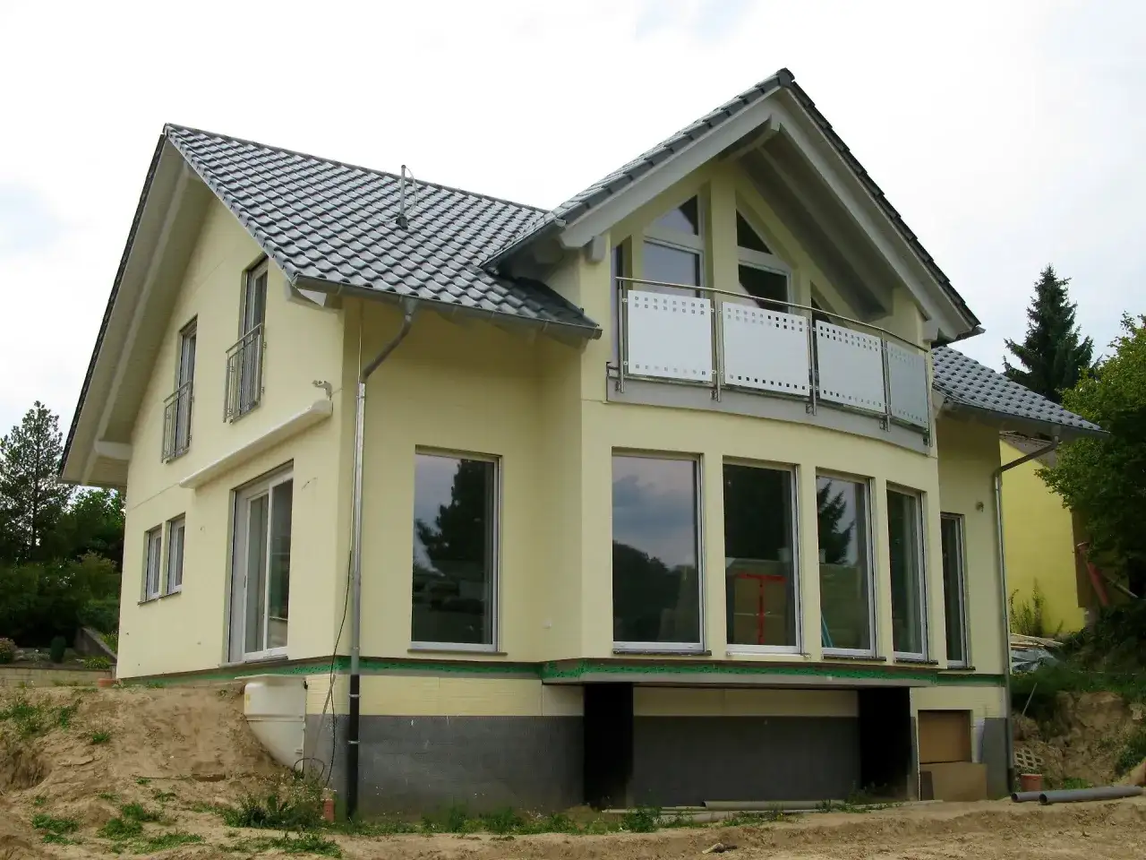 Nowy dom z żółtą elewacją i dachem w kształcie litery L. Duże okna i balkon dodają mu nowoczesnego charakteru.