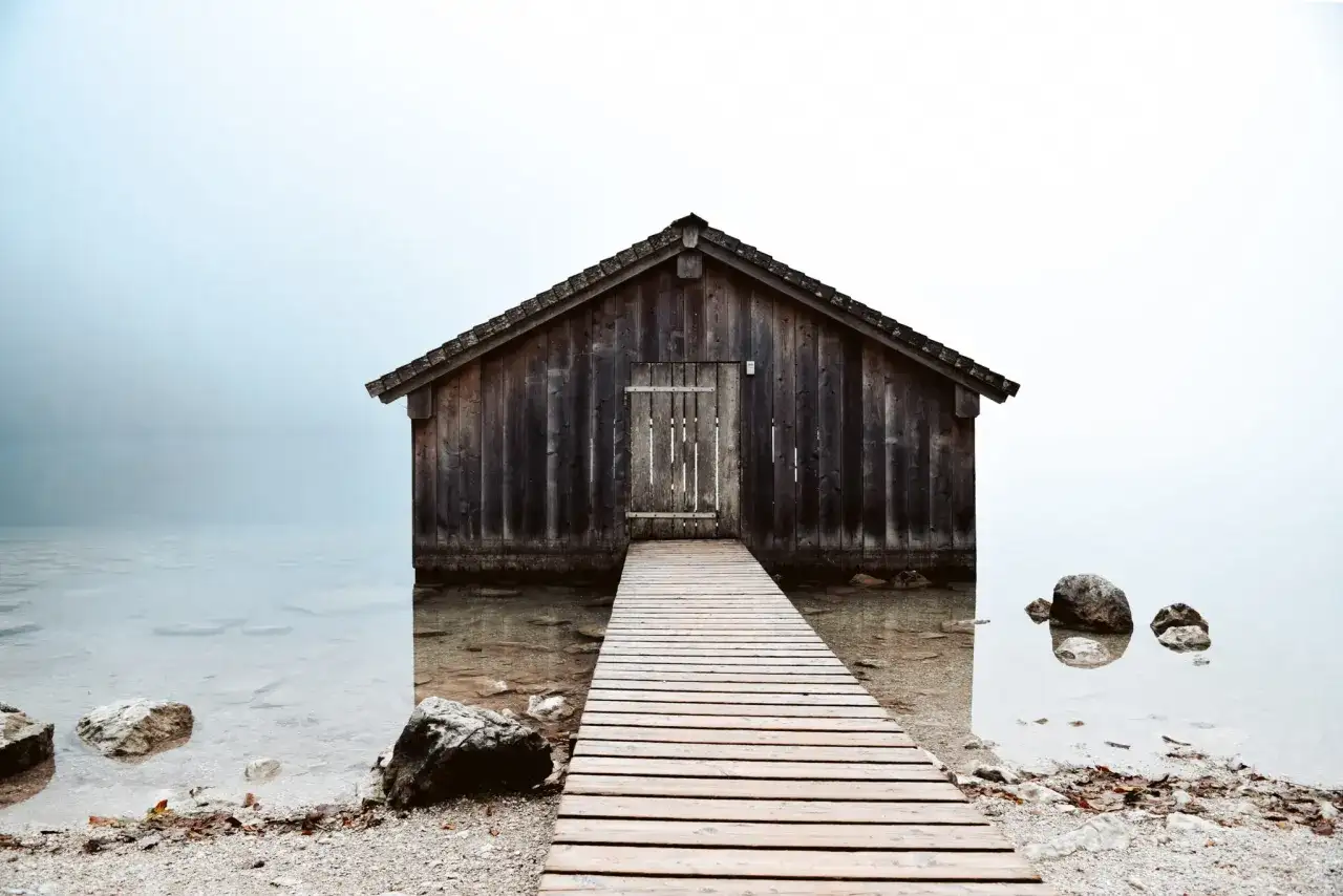 Drewniana szopa nad jeziorem, z pomostem prowadzącym do drzwi. Kompozycja otwarta przykłady.