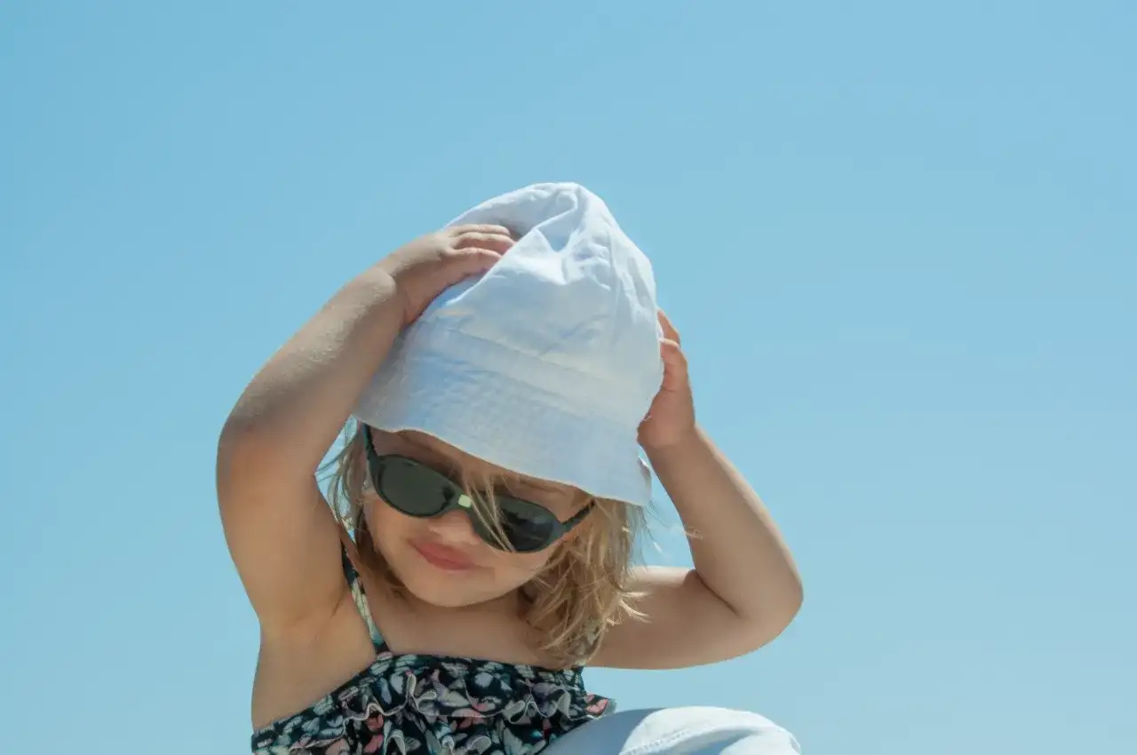 Dziecko w okularach przeciwsłonecznych i białej czapce UV dla dzieci, z rękami na głowie, na tle błękitnego nieba.