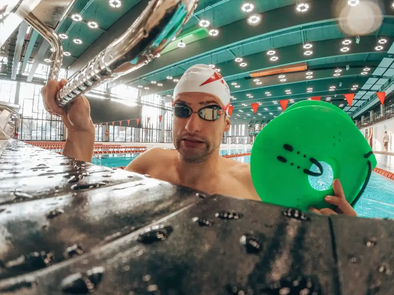 Pływak w czepku i okularach trzyma zielone łapki do pływania, gotowy do treningu.