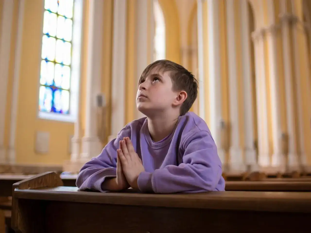 Chłopiec w fioletowej bluzie modli się w kościele, patrząc w górę.
