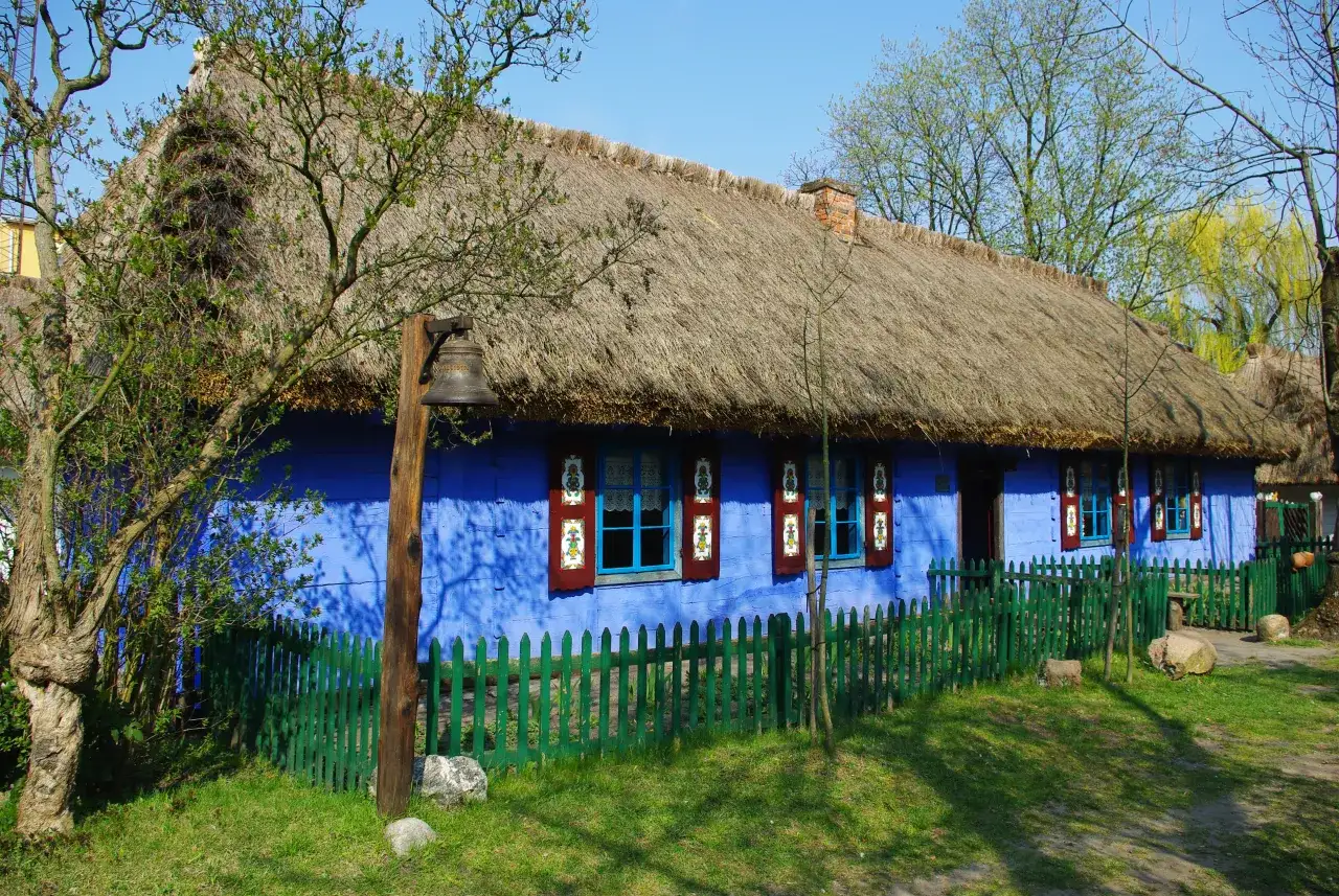 Niebieski dom ze słomianym dachem, zielonym płotem i dzwonkiem na drewnianym słupie.