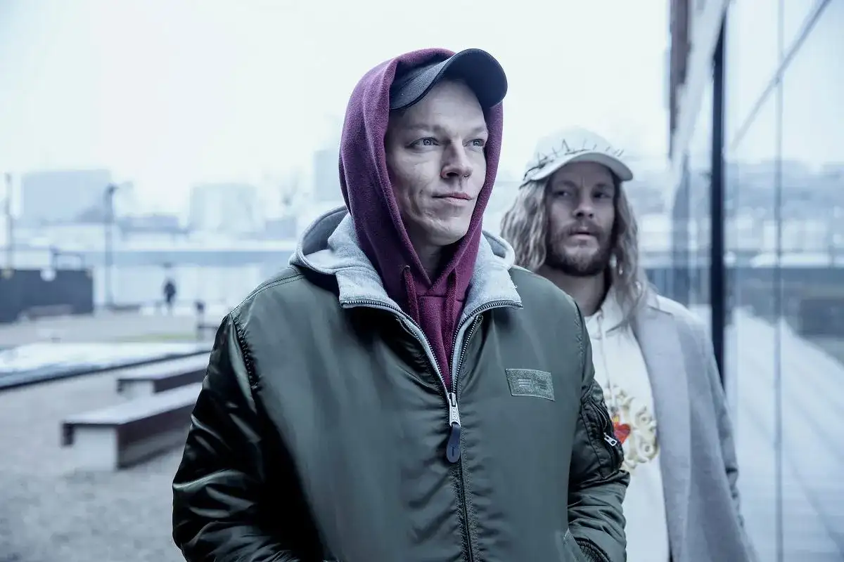 Dwóch mężczyzn w kapturach i czapkach, wyglądają jakby szli na jakieś seanse. Inni ludzie w tle.