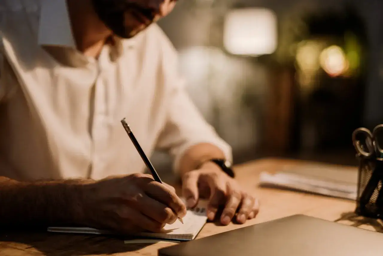 Mężczyzna w białej koszuli pisze ołówkiem w notatniku. Tworzy wartościowy branded content, pracując nad ważnym projektem.