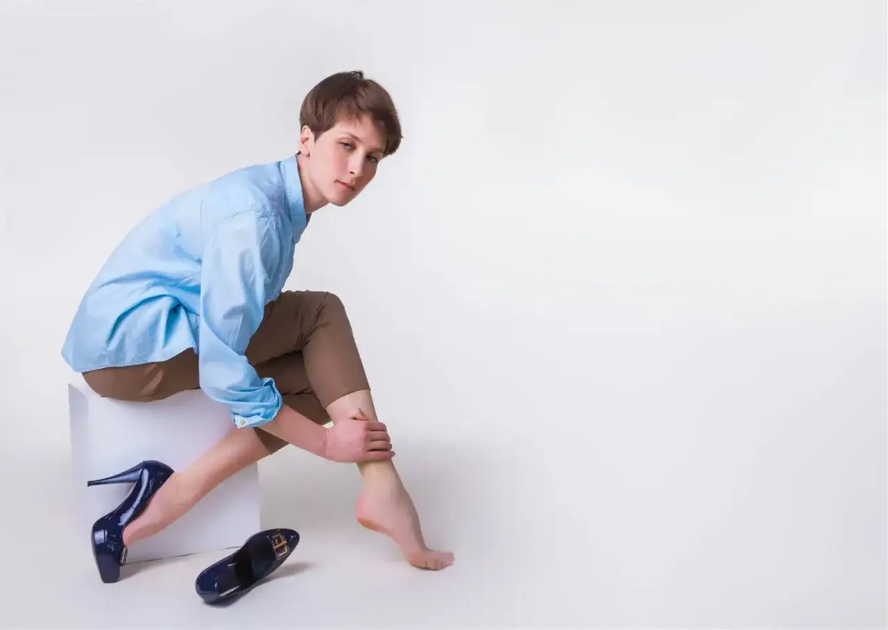 Kobieta w niebieskiej koszuli i brązowych spodniach masuje bolącą łydkę. Obok leżą wysokie obcasy, sugerując domowe sposoby na ból łydek.