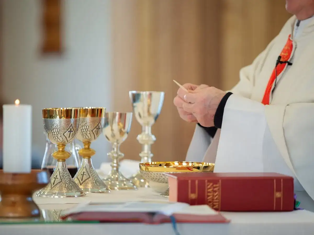 Ksiądz w białej szacie trzyma hostię, obok kielichy i mszał.