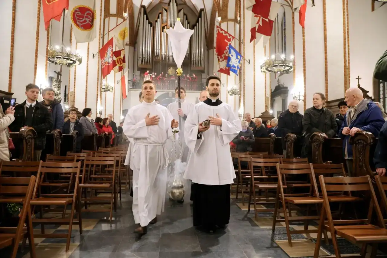 Liturgia Wielkiego Czwartku w kościele. Dwóch ministrantów w białych szatach niesie kadzidło i chorągiew.