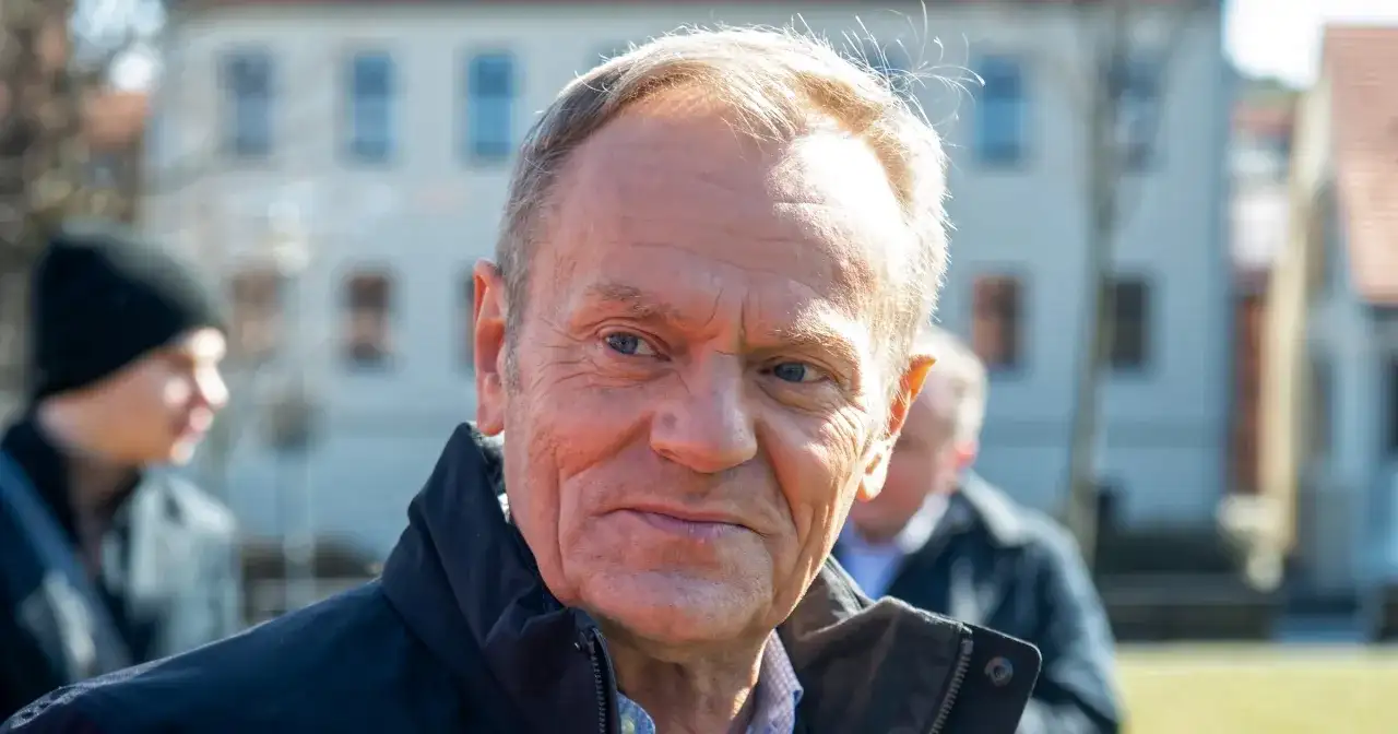 Portret Donalda Tuska w czarnej kurtce, z innymi osobami w tle.