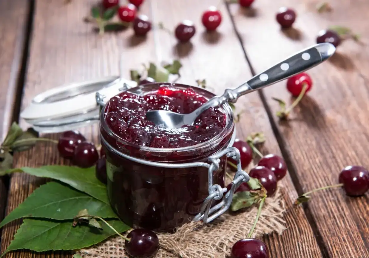 Słoik pełen domowego musu wiśniowego, z łyżeczką gotową do degustacji. Wokół świeże wiśnie i liście.
