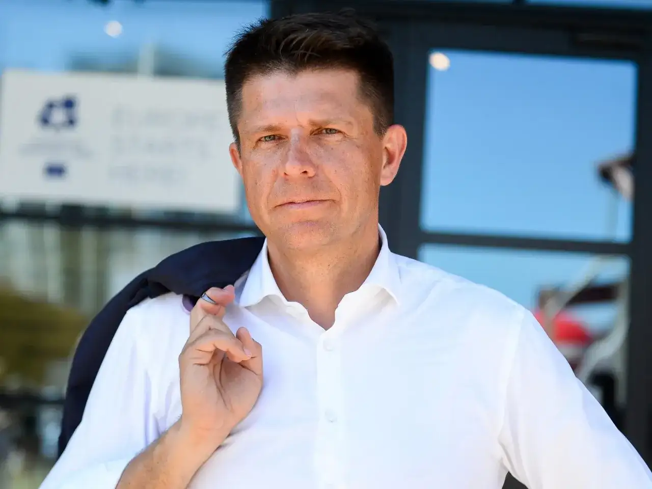 Ryszard Petru w białej koszuli, z marynarką na ramieniu, na tle budynku.