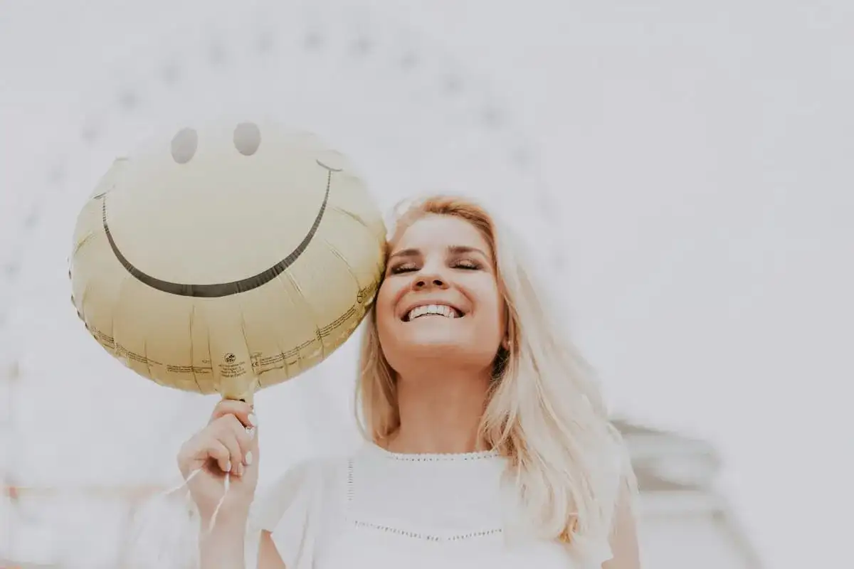 Uśmiechnięta blondynka trzymająca balon w kształcie buźki. Dobre żarty po angielsku poprawiają humor.
