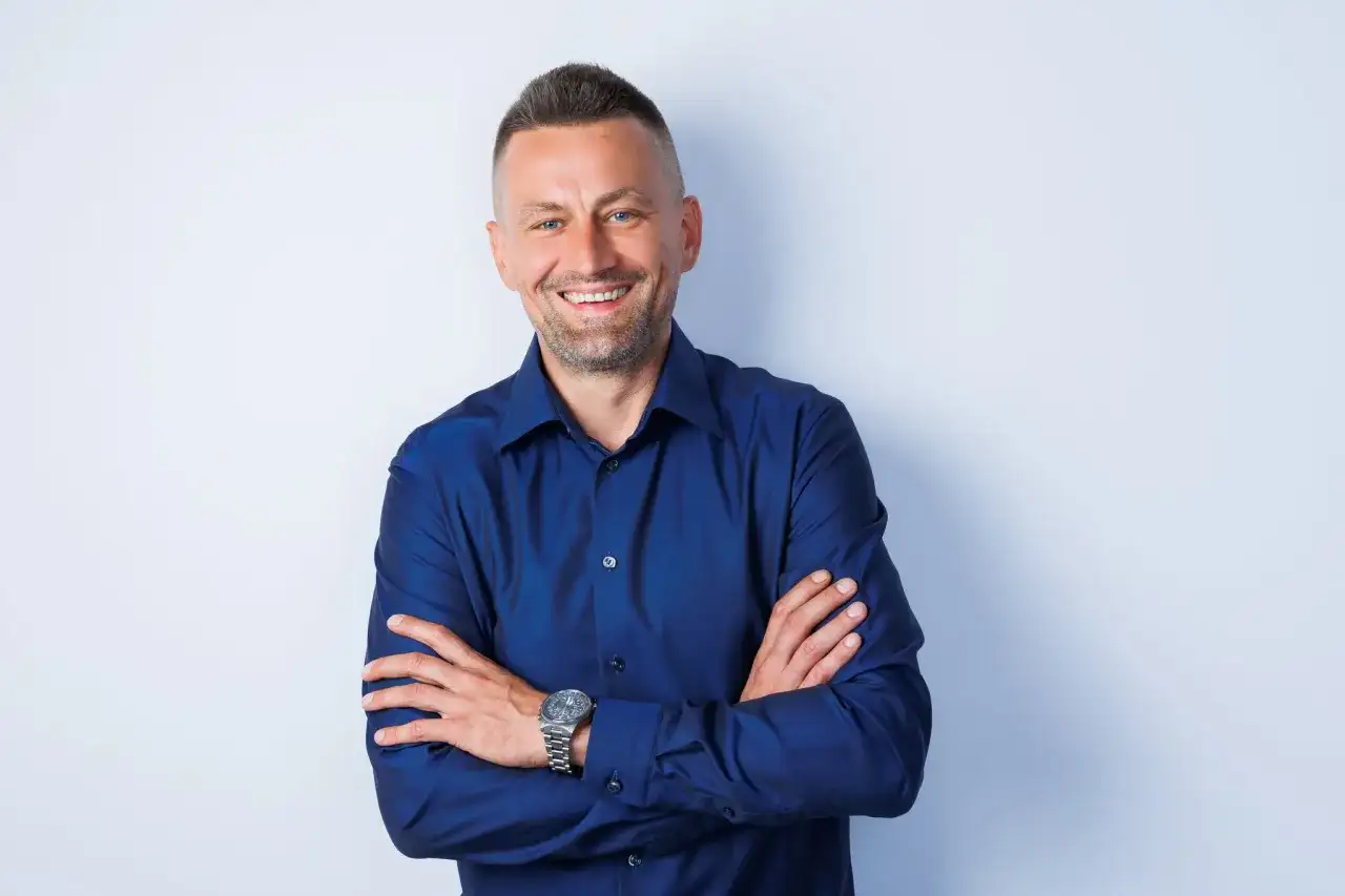 Uśmiechnięty mężczyzna w granatowej koszuli, z rękami skrzyżowanymi na piersi. Idealne zdjecie profilowe meskie.