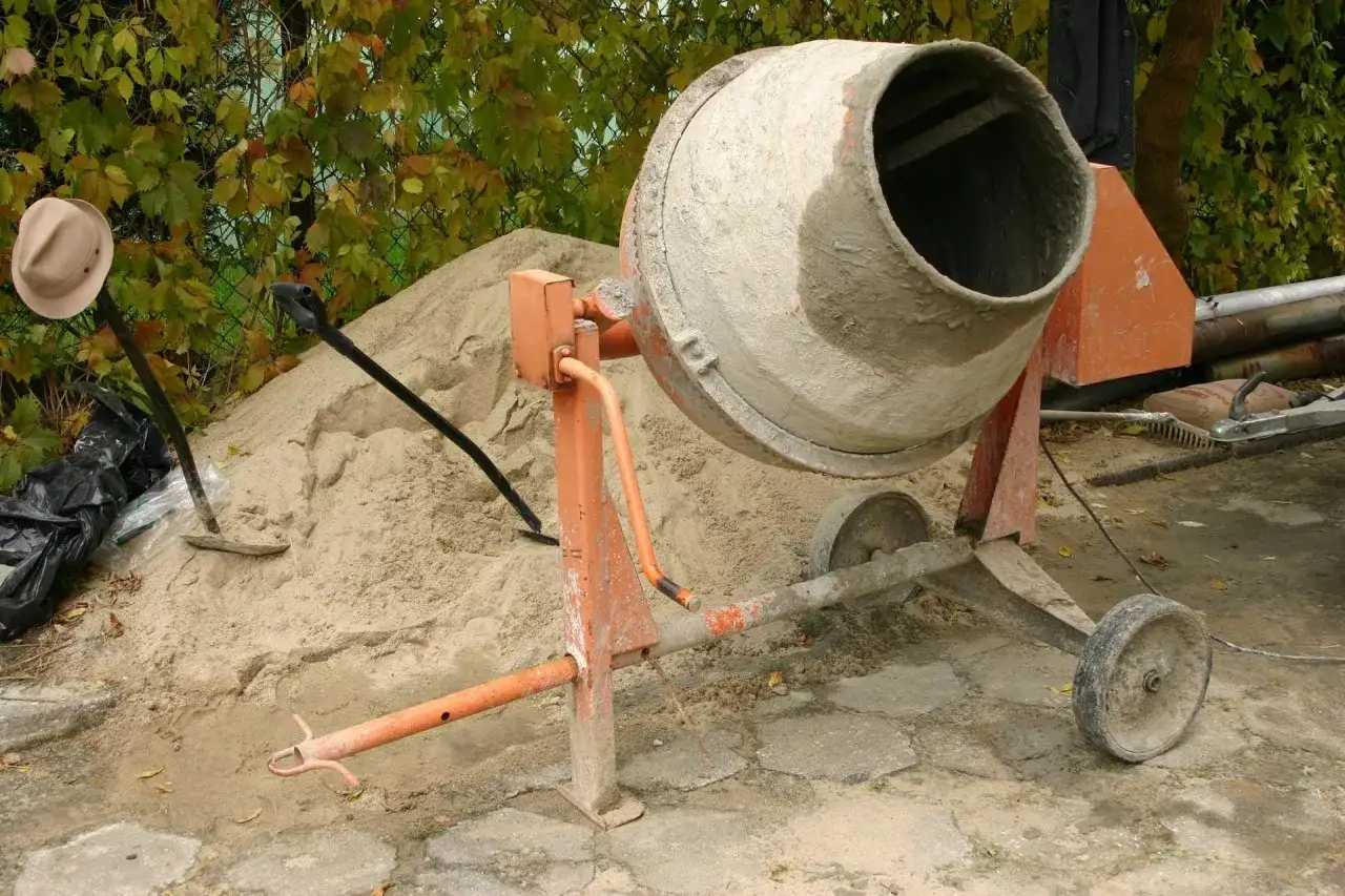 Betoniarka 120l czeka na cement. Obok piasek, łopata i kapelusz.