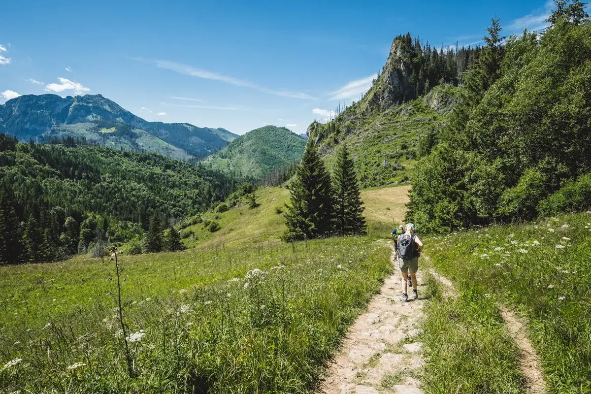 Turyści idą kamienistą ścieżką nad reglami, otoczeni zielonymi wzgórzami i skalistymi szczytami.