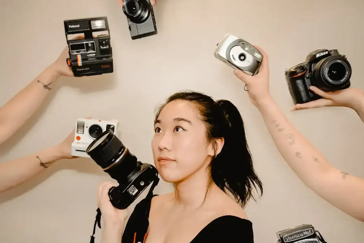 Kobieta z aparatem w ręku, otoczona przez inne aparaty: Polaroid, Nikon, cyfrówki. Idealna do wyboru, jaka lustrzanka na początek.