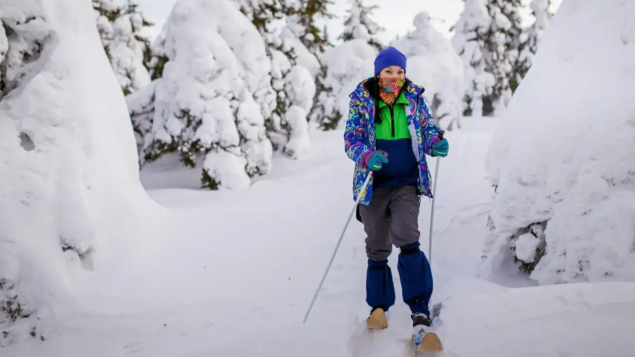 Kobieta w kolorowej kurtce i czapce narciarskiej na nartach w zaśnieżonym lesie.