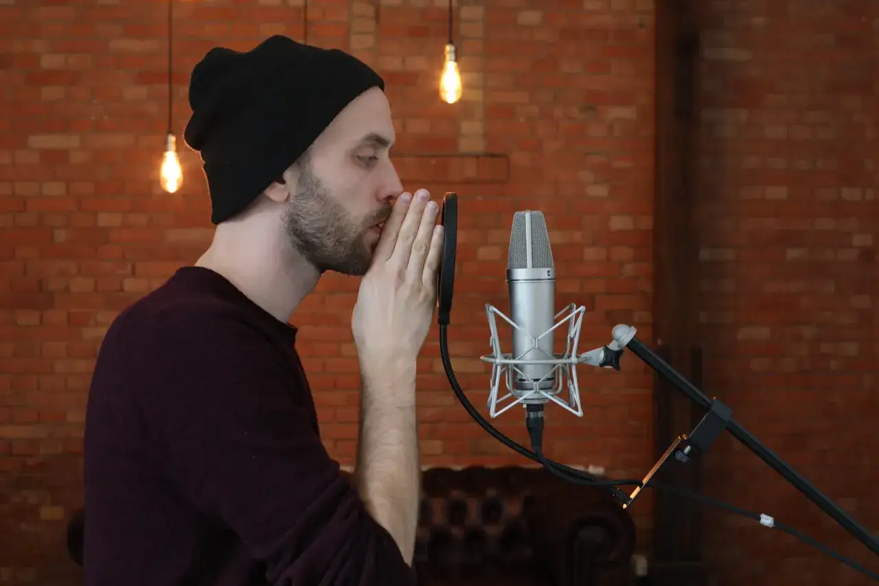 Mann mit schwarzer M&uuml;tze spricht ins Mikrofon, sucht nach rap ideen. Ziegelwand und Gl&uuml;hbirnen im Hintergrund.