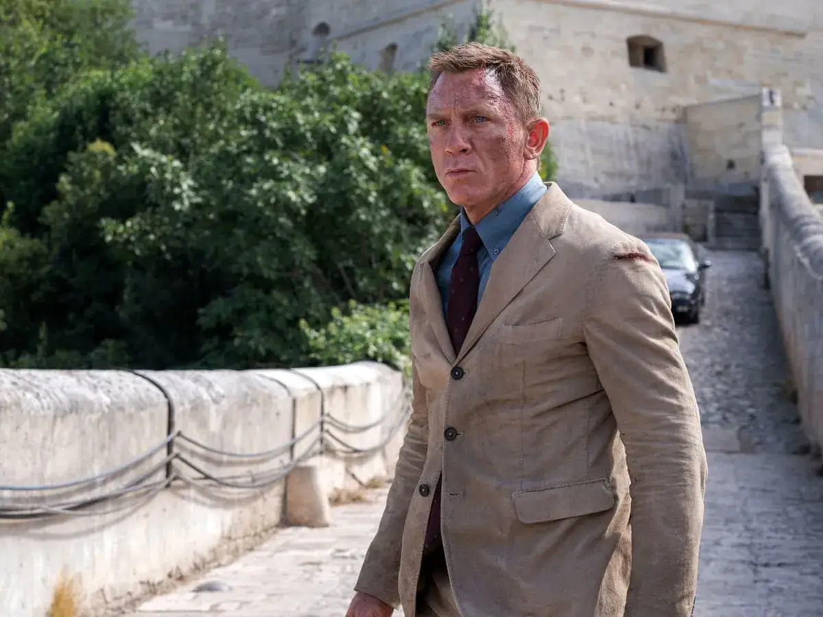 Ein Mann im James Bond Style, mit leicht zerrissener Kleidung, steht auf einem steinernen Weg vor einer alten Festung.