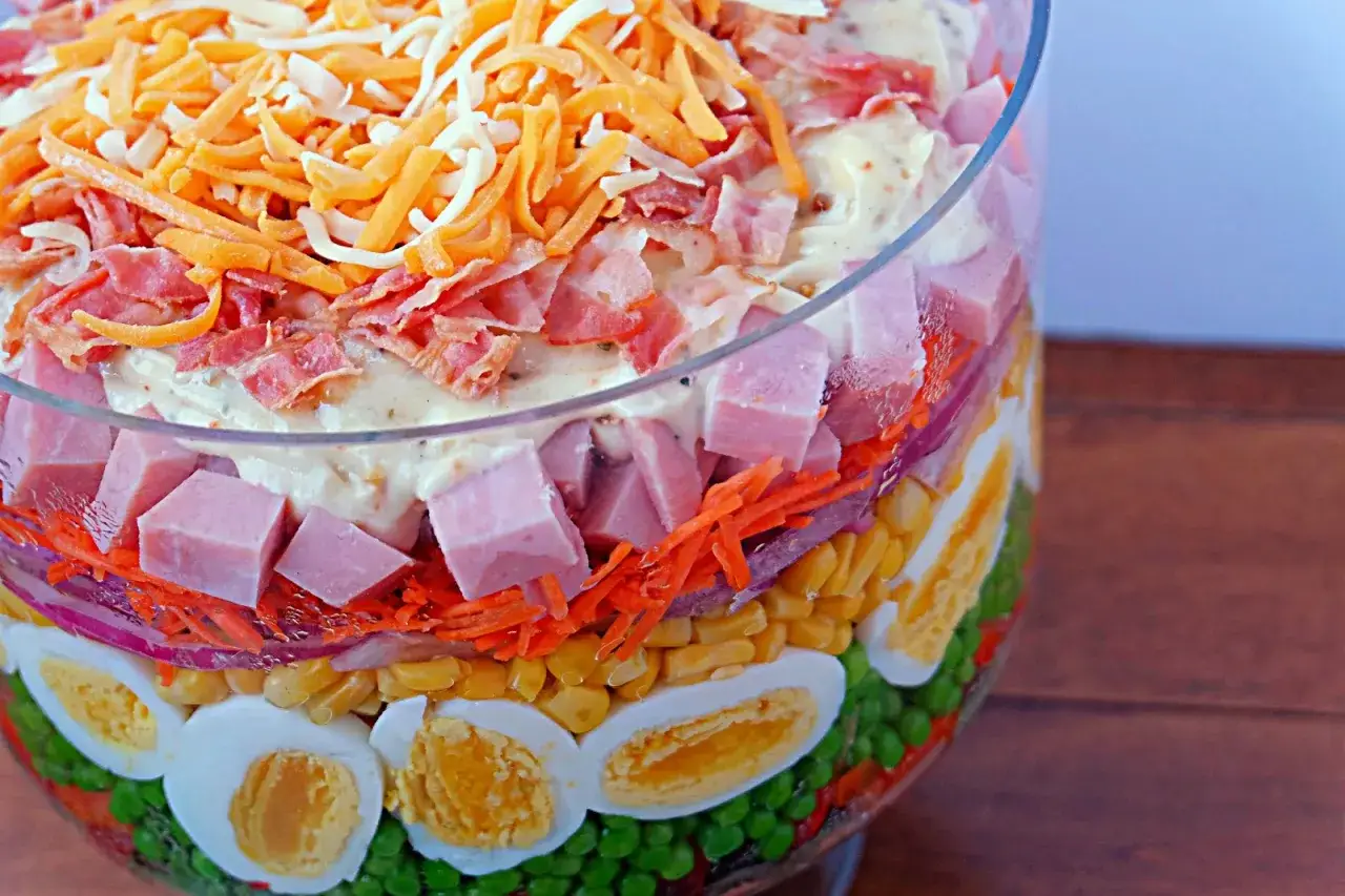 Une salade 7 étages colorée dans un bol en verre : petits pois, œufs durs, maïs, oignons rouges, carottes râpées, jambon en dés, puis une sauce crémeuse et du fromage râpé.