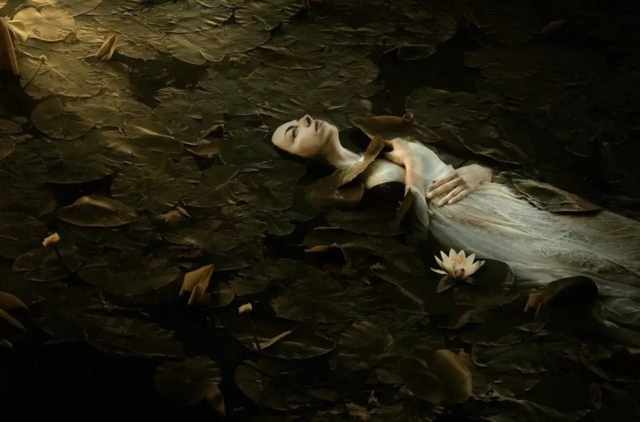 A woman in a white dress floats serenely on a bed of lily pads, a scene captured in evocative fine art photography.
